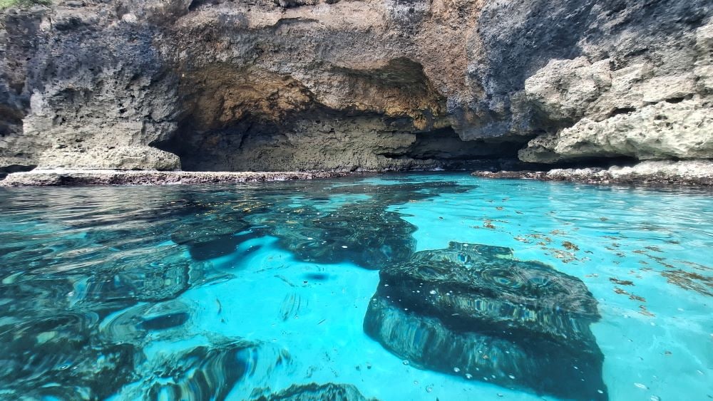The image captures a serene underwater landscape featuring large, textured rocks partially submerged in clear turquoise water. Sunlight filters through the surface, illuminating the scene and revealing plants swaying gently in the tide. A rocky cave entrance is visible in the background.