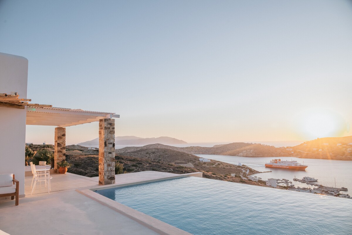 An infinity pool overlooks a stunning coastal landscape, with the sun setting over distant hills and water. A wooden terrace with a stone pillar structure offers seating for relaxation, enhancing the serene ambiance of the outdoor space.