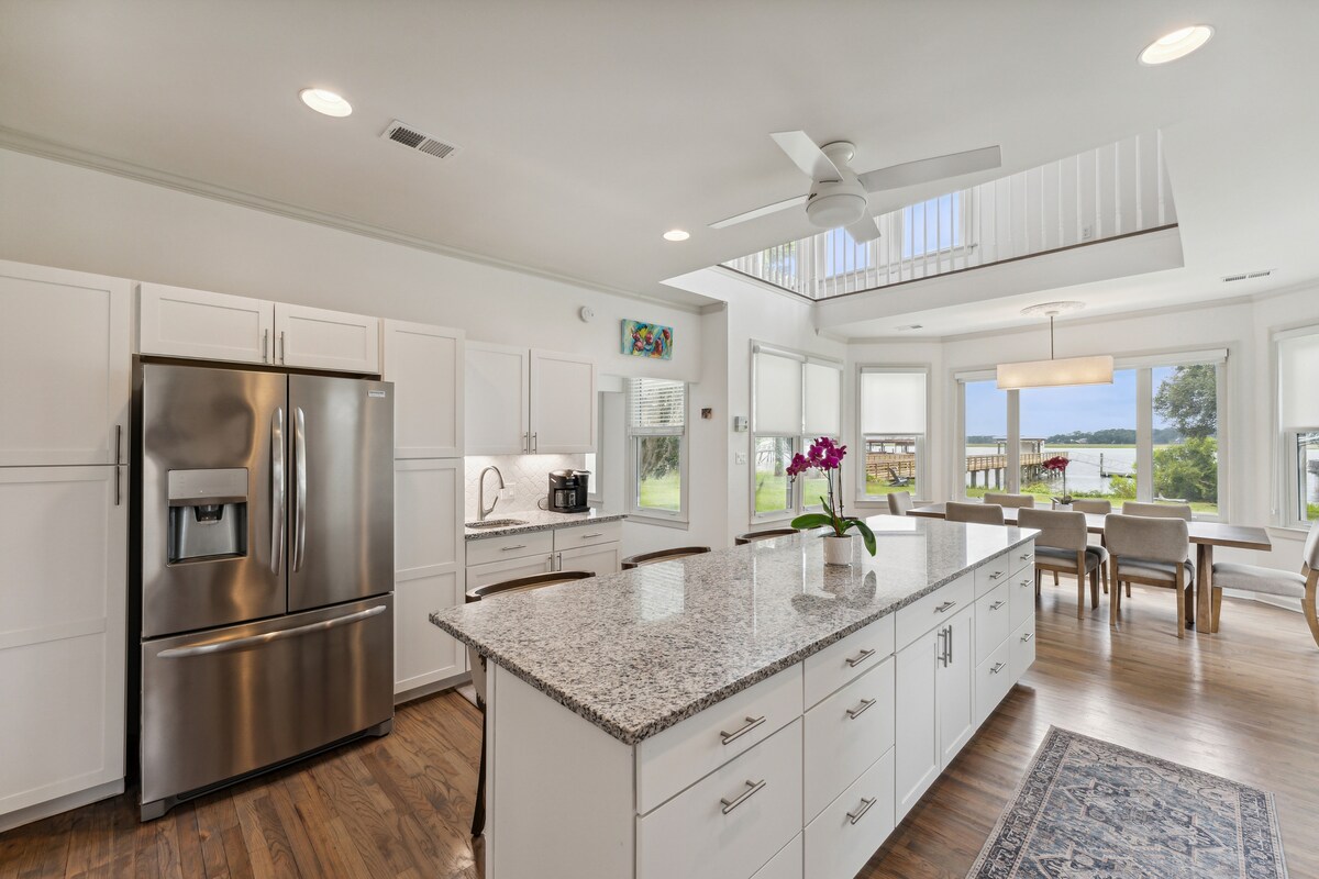An updated kitchen features a large island with granite countertops and modern stainless-steel appliances. Natural light floods the space through expansive windows, providing views of the waterfront and inviting an airy atmosphere. A dining area with a contemporary light fixture is visible in the background.