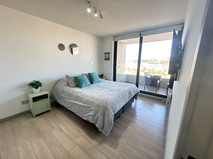 Habitación luminosa con cama matrimonial, ropa de cama en tonos cálidos, televisor frente a la cama y lámparas de noche. Su decoración natural y detalles acogedores crean un ambiente perfecto para descansar y relajarse junto al mar.