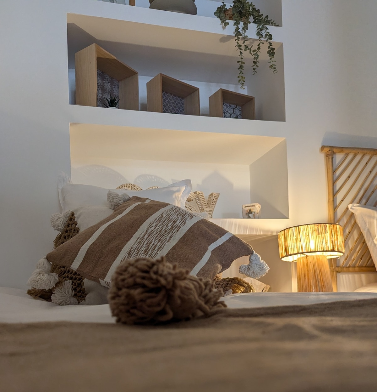 A cozy sleeping area features a spacious bed adorned with decorative pillows. A decorative shelf is positioned above, showcasing geometric wooden pieces and greenery. A soft bedside lamp casts a warm glow, providing an inviting atmosphere.