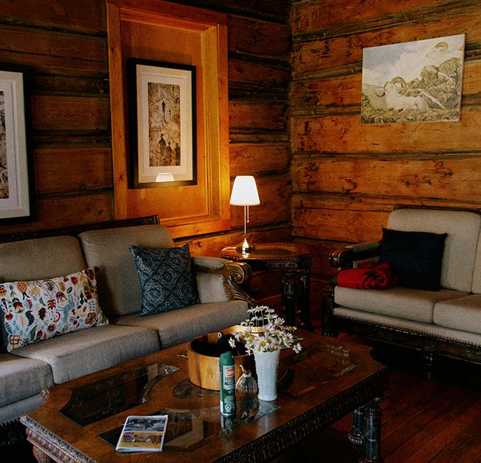 A cozy sitting area features a log cabin wall, with two sofas adorned with decorative pillows. A low wooden coffee table is centered, holding a vase of flowers and a magazine. Soft light is provided by a lamp, creating a warm ambiance.