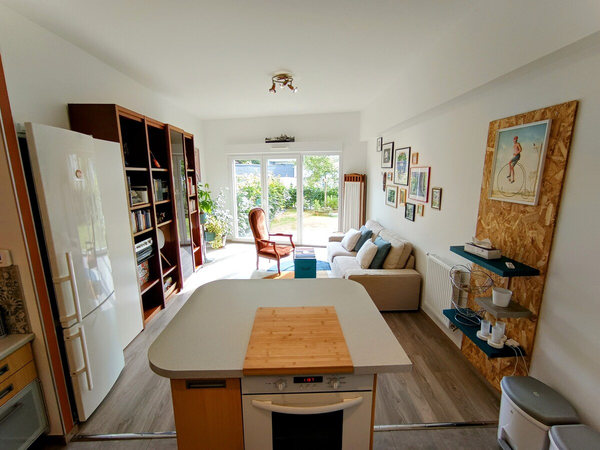 A modern kitchen and living area is presented, featuring a central countertop with a wood surface. A comfortable chair faces a sofa, accompanied by natural light from large sliding doors opening to a garden. Bookshelves and wall art add visual interest to the space.