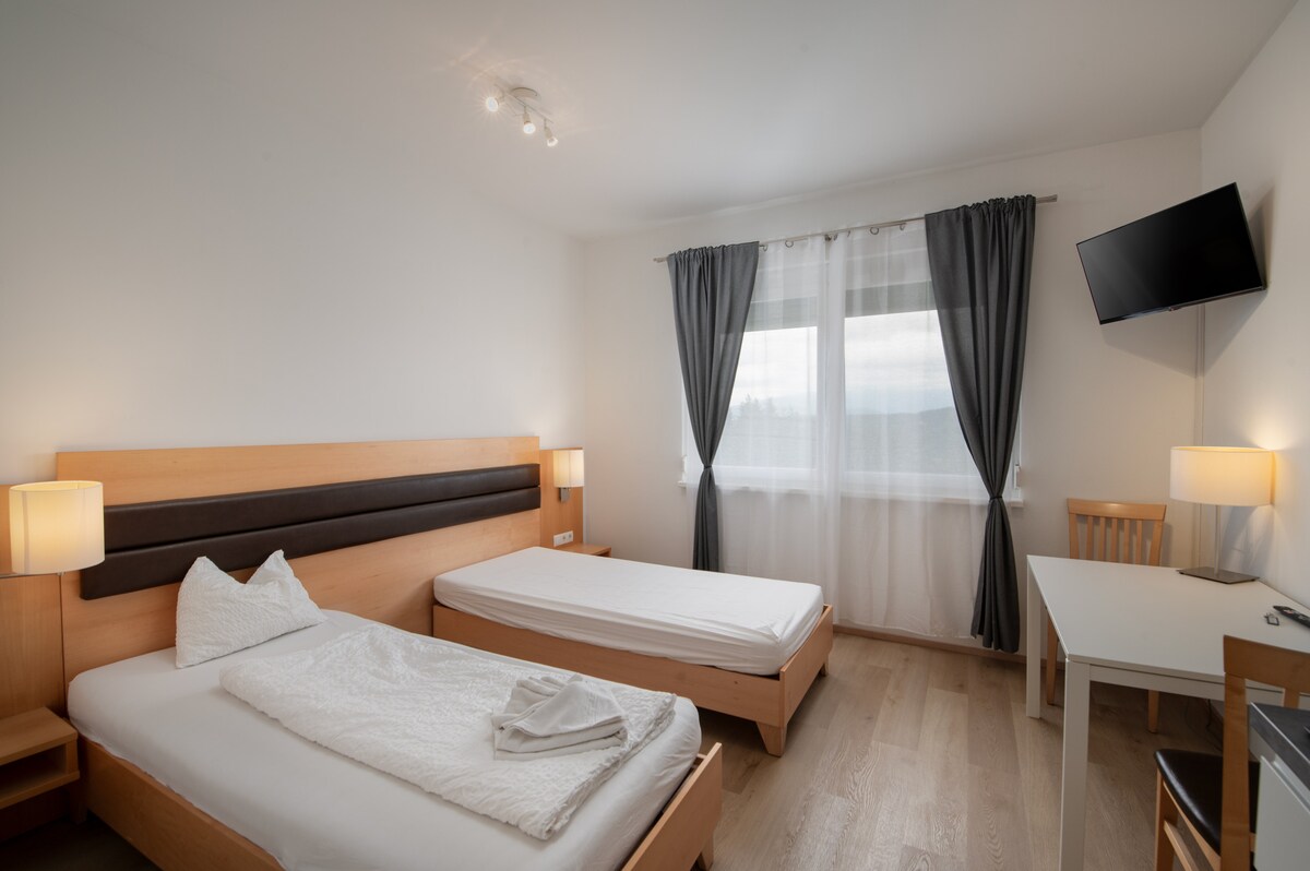 A well-lit bedroom features two beds, one made with a white blanket and the other prepared with fresh bedding. Soft, natural light enters through a large window, complemented by light curtains. A small desk and a wall-mounted TV are visible, providing a functional space.