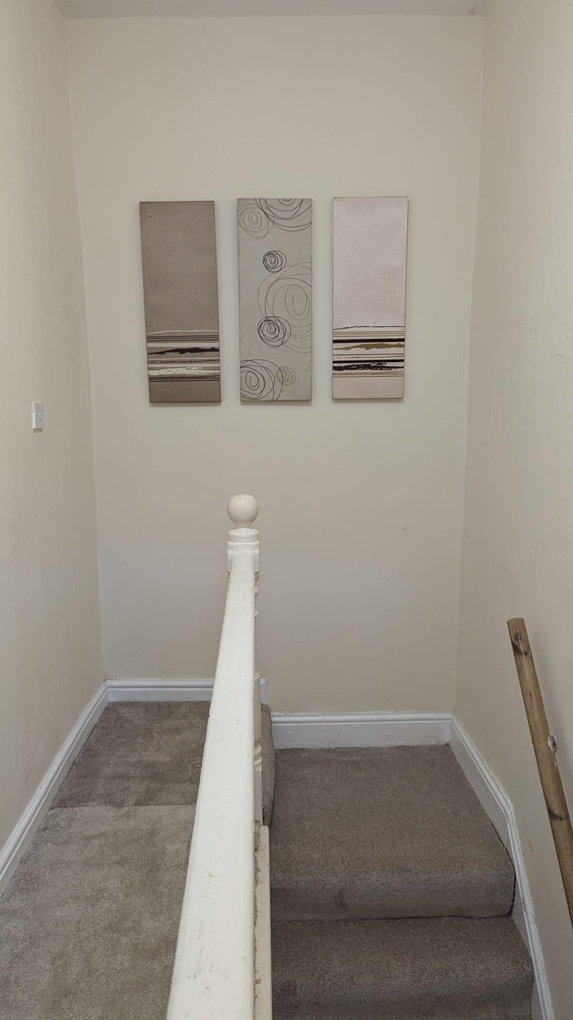 A stairway is shown leading upward, with soft beige walls and a carpeted floor. Three abstract canvas artworks are hung on the wall, featuring neutral tones that complement the surroundings. A white handrail provides safety and guidance along the stairs.