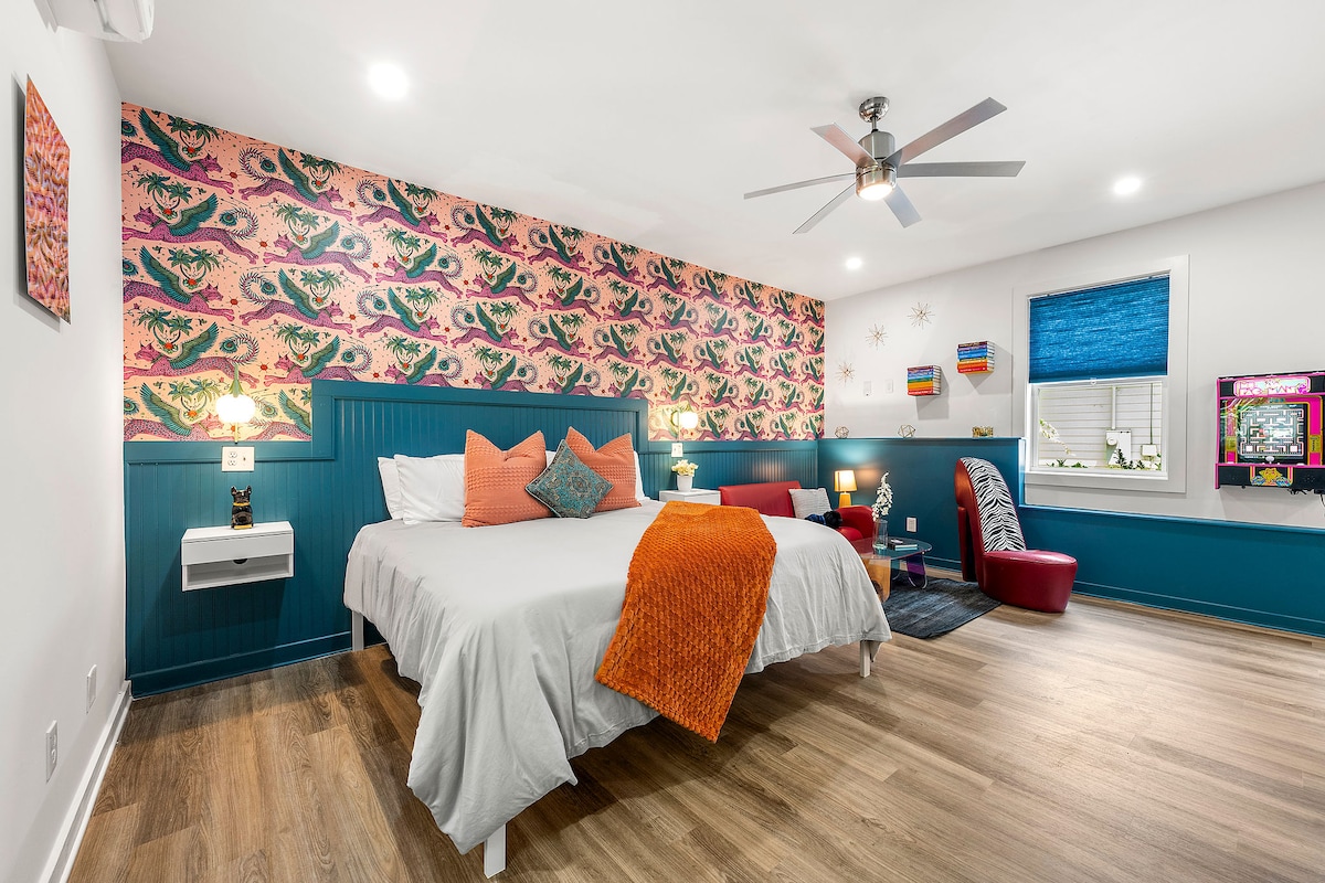 A studio layout features a spacious bed dressed in crisp linens with a vibrant orange throw. A colorful accent wall presents a whimsical pattern. The room includes a comfortable armchair and a Miss Pacman arcade game, with a ceiling fan providing airflow.