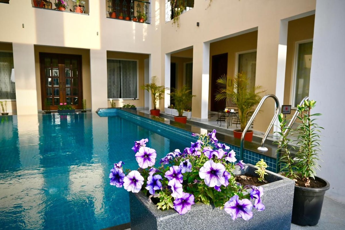 An indoor pool is featured, surrounded by elegantly designed rooms with large openings. Potted plants and vibrant purple flowers enhance the tranquil setting, creating a soothing atmosphere. Light reflects off the water, adding to the serenity of the space.