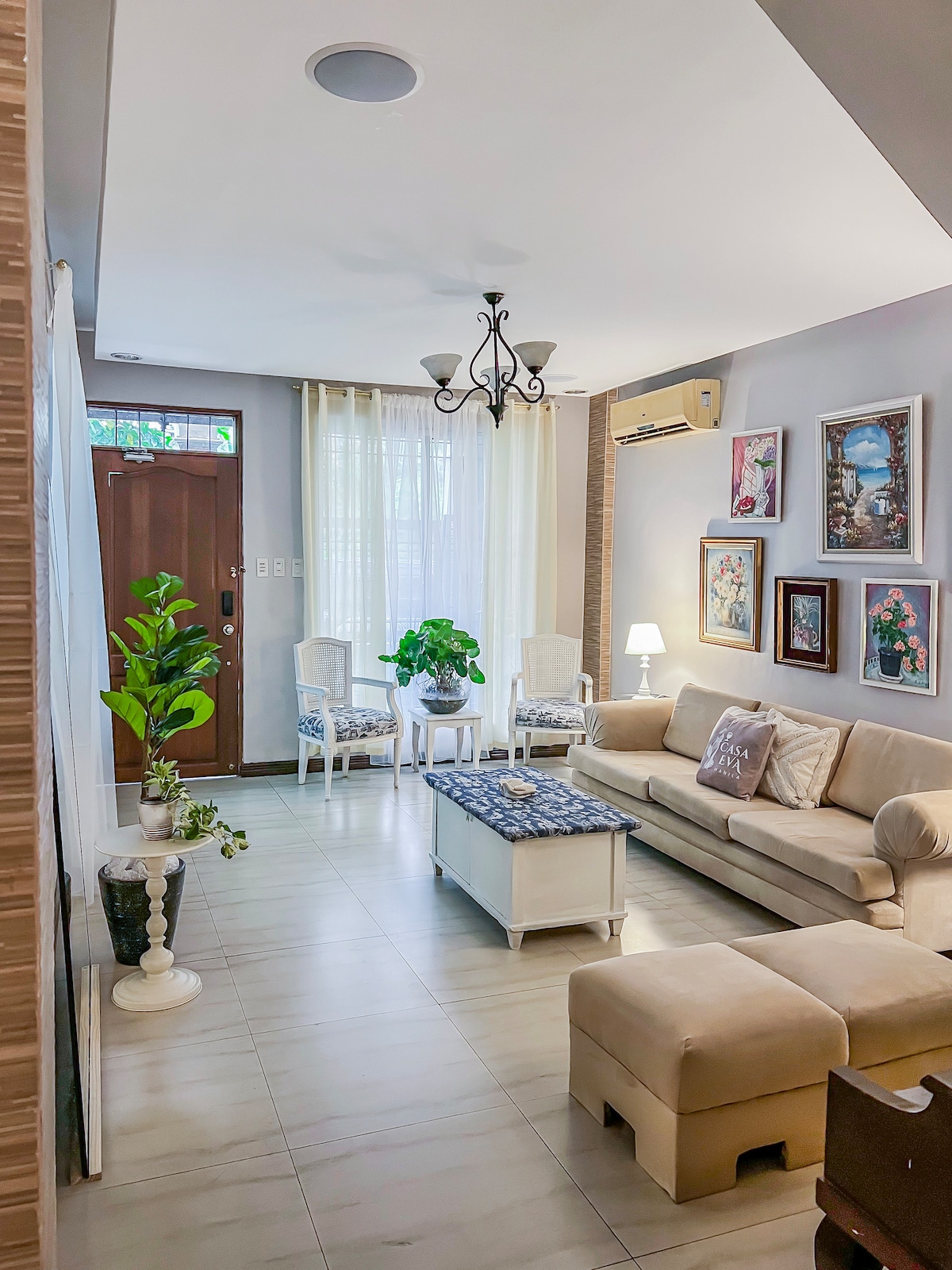 A cozy living room is presented with a light-colored sofa and an accent chair, accompanied by a coffee table. Natural light filters through sheer curtains, highlighting potted plants and wall art. A chandelier hangs overhead, adding a touch of elegance to the inviting space.