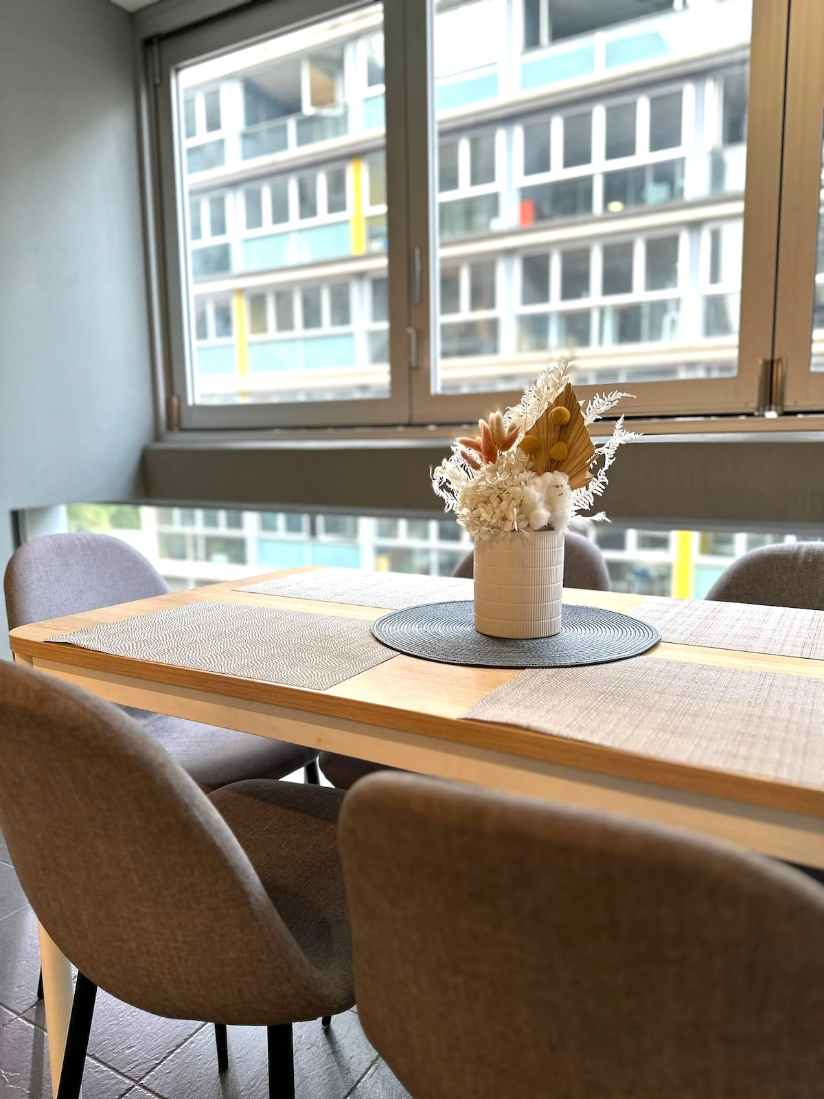 A dining area is presented with a wooden table surrounded by four upholstered chairs. A decorative vase filled with dried flowers is positioned at the center atop a placemat. Large windows provide natural light and a view of the building's exterior features.