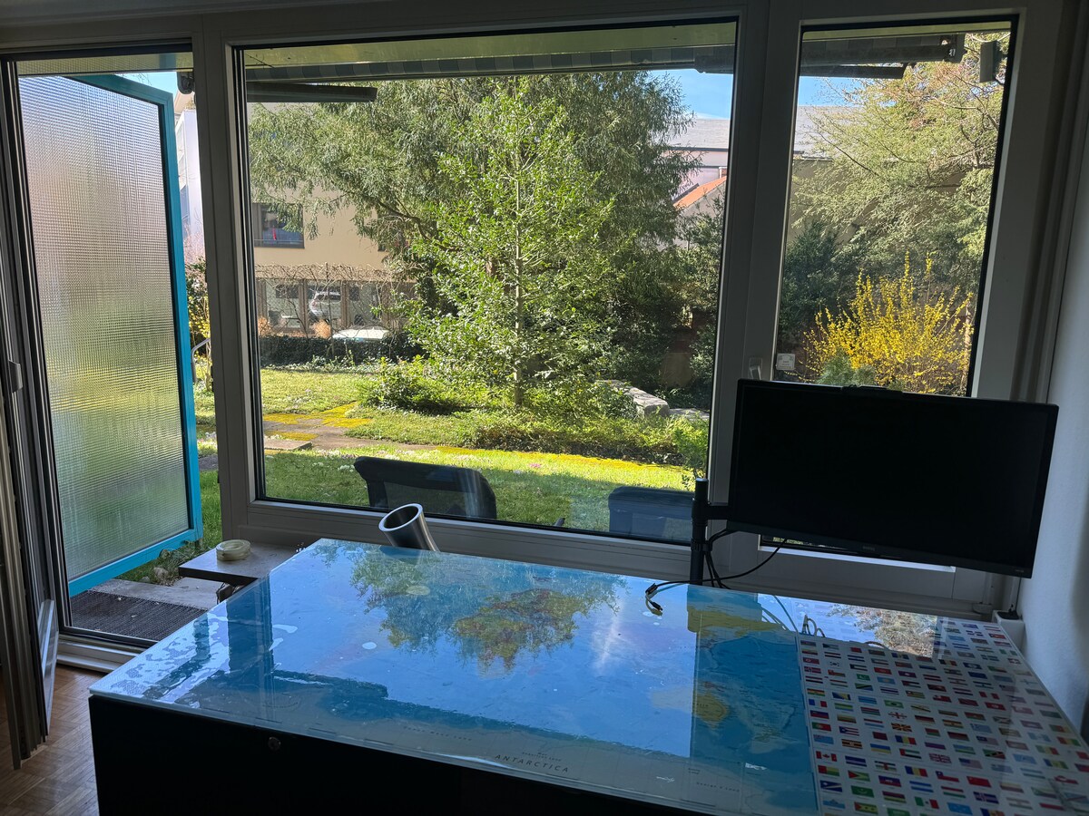 A workspace is visible with a large desk featuring a world map on the surface and a monitor positioned nearby. Through spacious windows, a lush green backyard is seen, complete with trees and seating areas, providing a serene outdoor view.