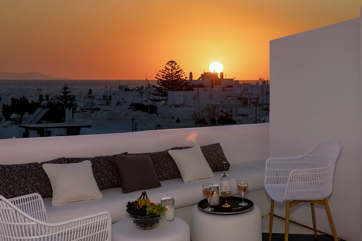 A shared rooftop terrace is seen during sunset, featuring comfortable seating arrangements. A round table is set with glasses of wine and a fruit platter, while the horizon displays warm tones as the sun sets over the Aegean Sea.