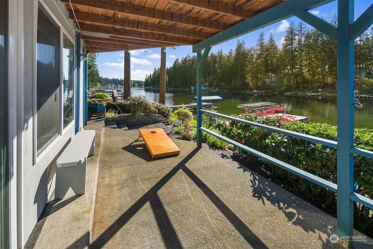 A covered patio area features a concrete floor and a wooden beam ceiling, extending towards the waterfront. A cornhole game set is positioned on the patio, surrounded by colorful flowers and greenery. The tranquil lake and trees are visible in the background.