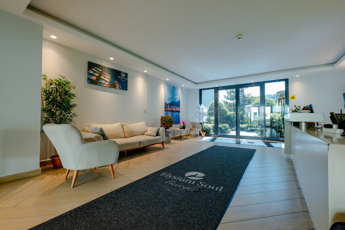 A spacious lobby area features a light-toned floor and stylish seating arrangements. A large welcome mat displays the name of the residence. Floor-to-ceiling glass doors allow natural light to fill the space and provide views of the outdoor greenery.