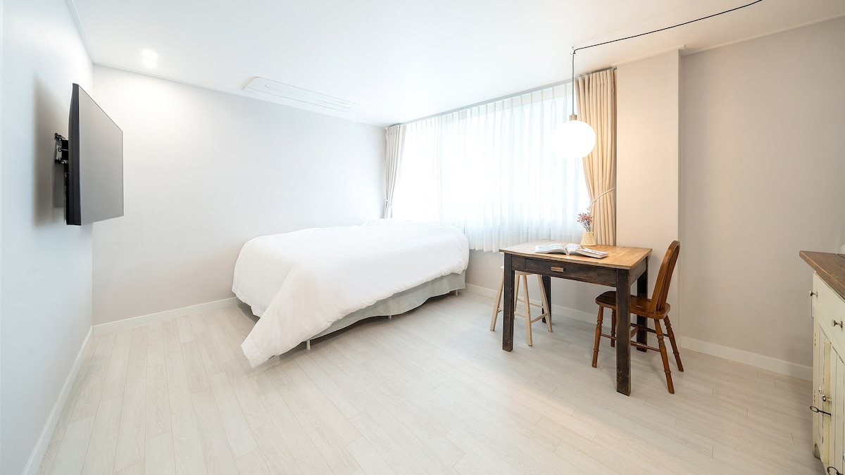 A spacious room features a large bed with crisp white bedding. A wooden table with two chairs is positioned near a window draped with light curtains. A wall-mounted television adds to the modern decor.