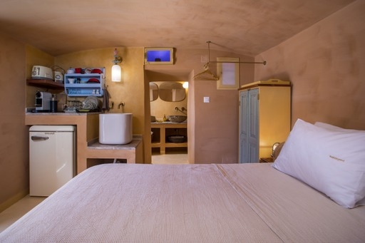 An earthy-toned studio space features a spacious bed and a small kitchenette in one corner. A sleek bathroom area is visible, complemented by an open design. Soft lighting is provided by ceiling fixtures, and a wardrobe stands next to the bed, enhancing the room's functionality.