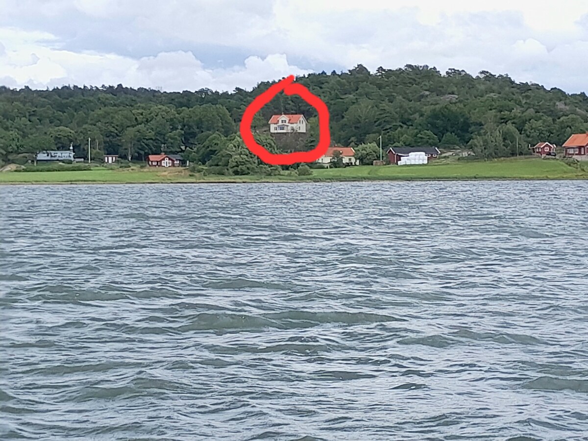 The image captures a distant view of a house set atop a hill, surrounded by lush greenery. The structure features a red-roofed top and is positioned above various smaller buildings visible along the shoreline. Water reflects the sky above, lending depth to the scene.