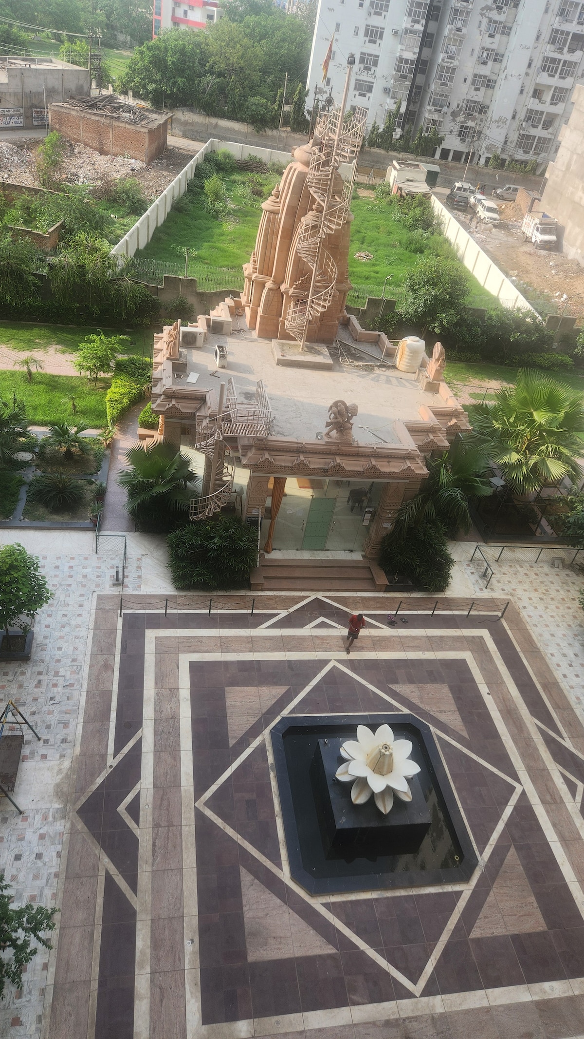 An aerial view captures the society mandir adorned with intricate carvings and a prominent spire. A central fountain featuring a white lotus is surrounded by landscaped greenery and decorative tile work, creating a serene focal point in the outdoor space.