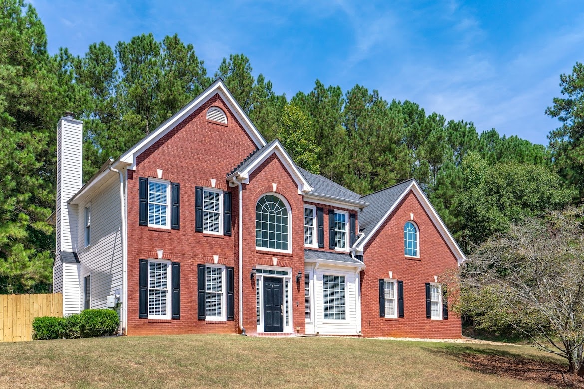 A two-story brick house is set against a backdrop of lush green trees. The symmetrical façade features a large arch window above the front entrance, flanked by smaller windows in a traditional style. A well-maintained lawn complements the inviting exterior.