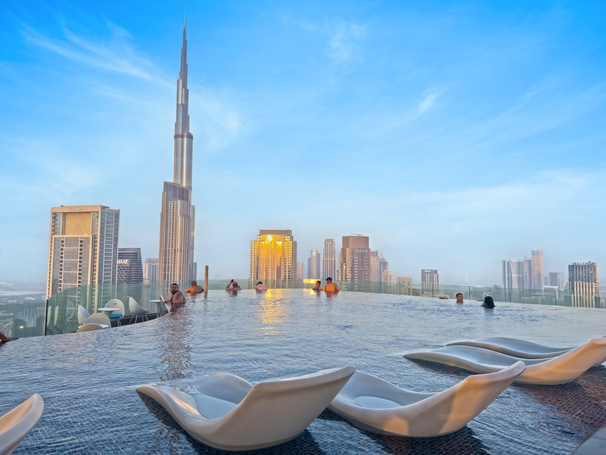 An infinity pool is positioned at a height, providing panoramic views of the Dubai skyline, including the iconic Burj Khalifa. Comfortable lounge chairs are set along the edge of the pool, creating a serene spot to relax while enjoying the stunning surroundings.