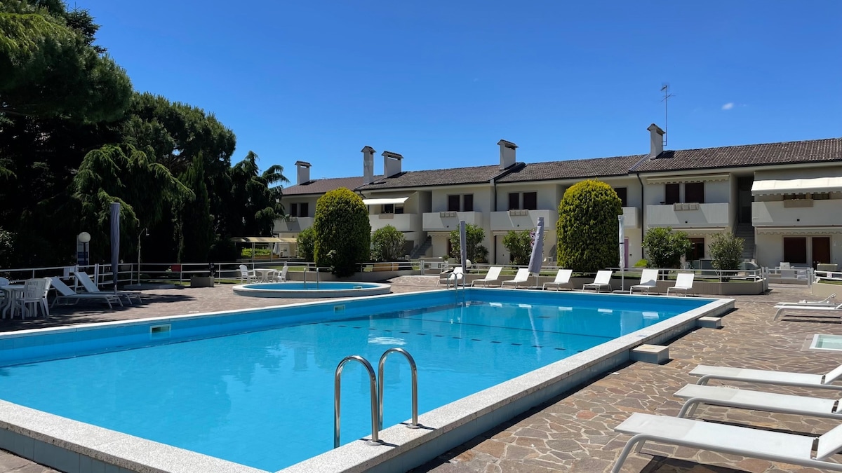 The image shows a clear, inviting swimming pool surrounded by a paved area with lounge chairs. In the background, two-story buildings with balconies are present, framed by lush greenery. The bright blue sky complements the tranquil atmosphere of the outdoor space.
