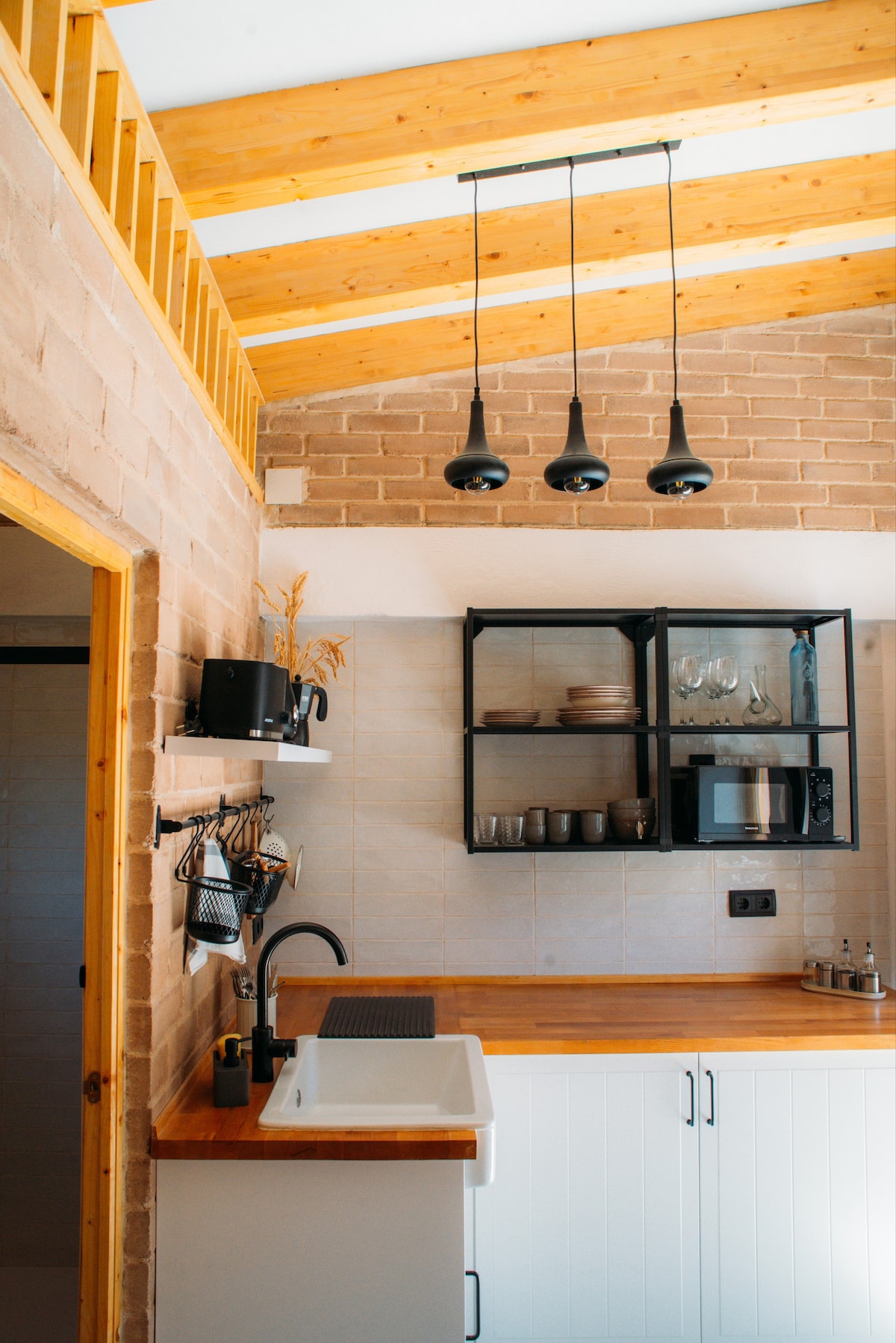 The kitchen features wooden cabinetry and a rustic brick wall, with a sink and countertop for meal preparation. Glass shelves hold dishware and glasses, while a microwave is visible. Three pendant lights hang from the ceiling, illuminating the space.