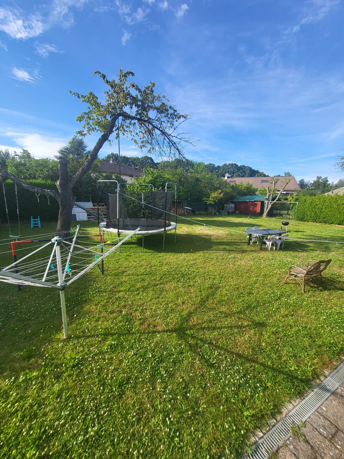 A spacious garden area features a trampoline, a slide, and a swing set among green grass. Outdoor seating is provided with a table and chairs. Trees offer shade, and a clothesline is visible near the garden edge, enhancing the outdoor space for relaxation and play.