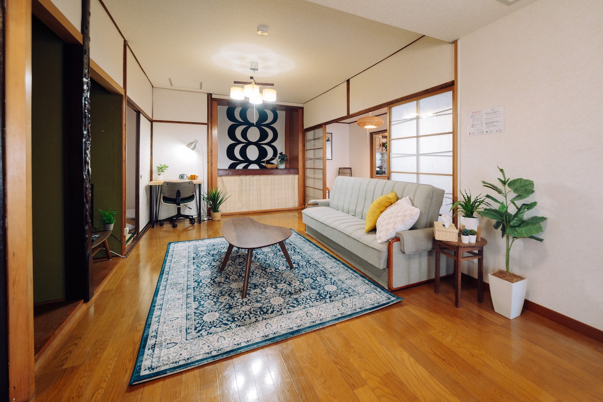 A spacious living area features a green sofa adorned with decorative pillows, positioned beside a round wooden coffee table. A vibrant area rug adds warmth to the polished wooden floor, with potted plants accentuating the space. A work desk and chair are visible in the background.