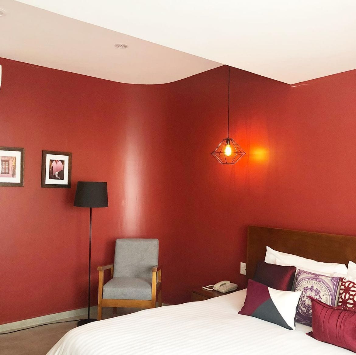 A room features deep red walls that create a warm ambiance, complemented by a spacious bed adorned with colorful pillows. A modern pendant light hangs above, while a gray armchair sits beside a floor lamp, providing a cozy reading nook. Framed artwork decorates one wall.