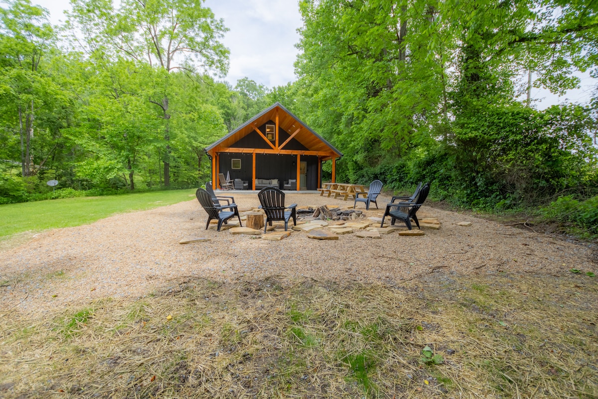 A welcoming outdoor space features a fire pit surrounded by several black Adirondack chairs. Gravel underfoot creates a rustic feel, while greenery envelops the area, providing a serene backdrop. The cabin's wooden exterior and porch are partially visible, adding to the inviting atmosphere.