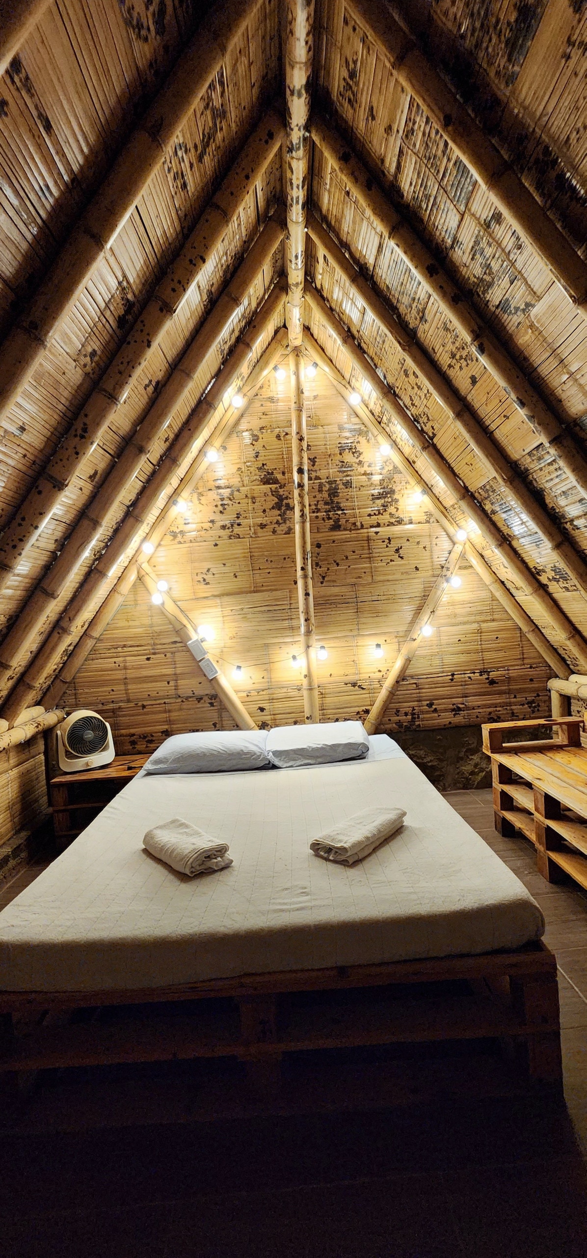 An attic room features a spacious bed with white linens and rolled towels. The wooden walls are complemented by warm, ambient lighting from hanging bulbs, creating a rustic atmosphere. A small fan and wooden furniture provide essential amenities in this inviting space.