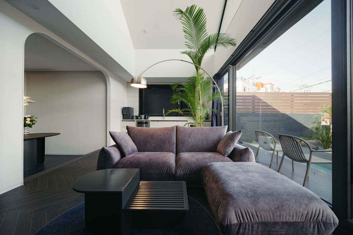 A modern living area features a plush, curved sectional sofa and a sleek coffee table. A large glass door allows natural light to fill the space, overlooking an outdoor area. Potted plants add a touch of greenery, creating a calming environment.