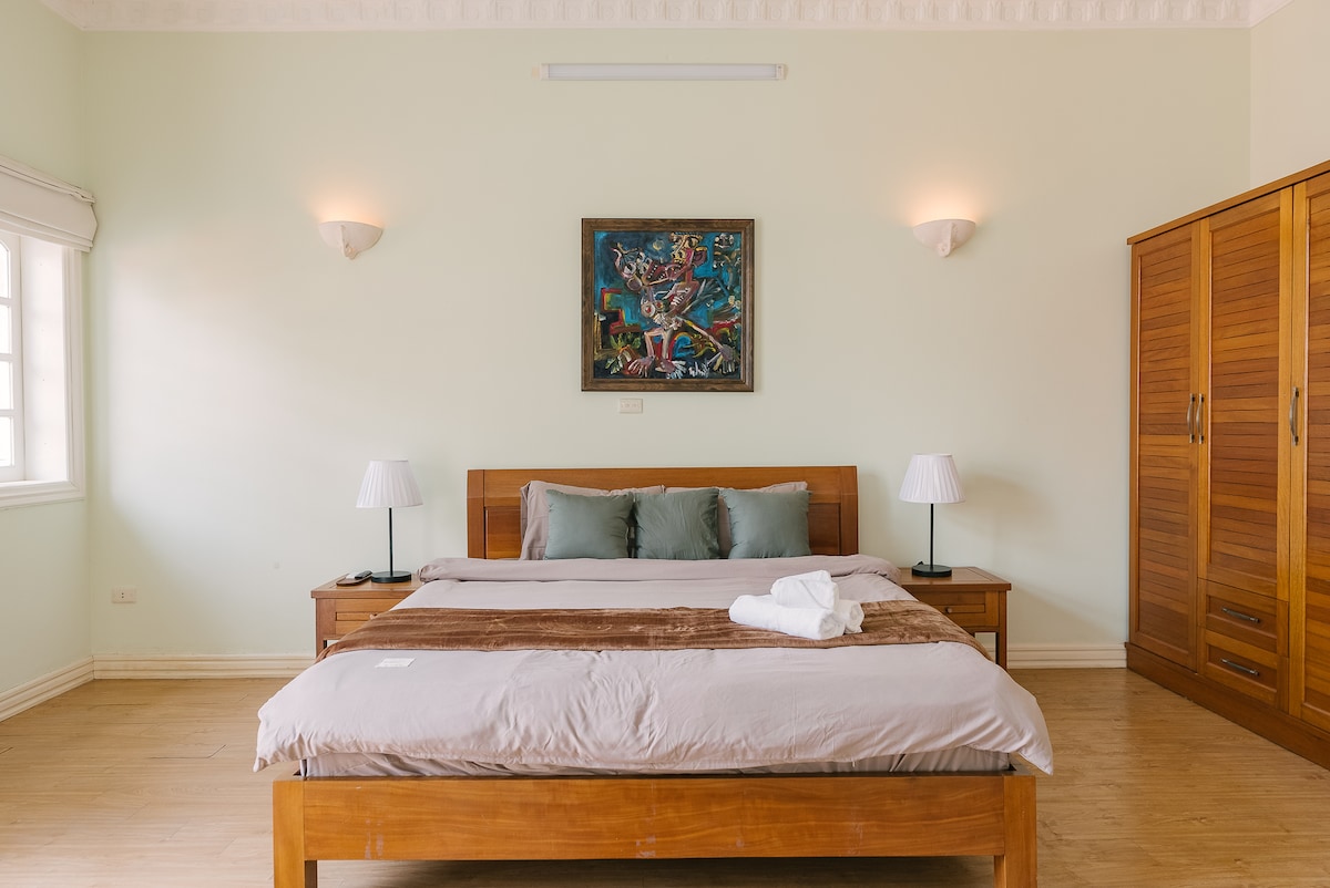 A spacious bedroom features a large bed with neutral-toned linens and decorative pillows. Two bedside lamps are placed on matching wooden tables. A wardrobe occupies one corner, while a vibrant piece of artwork adorns the wall above the headboard, enhancing the room's character.