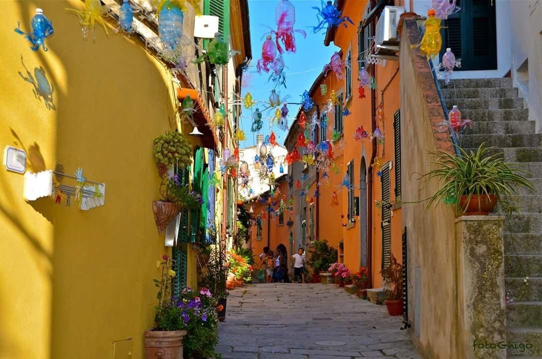 A narrow, charming street is lined with colorful buildings featuring vibrant yellow and orange facades. Decorative lanterns hang overhead, adding a festive touch. Potted plants and flowers adorn the edges, while groups of people can be seen walking along the cobblestone pathway, creating a lively atmosphere.