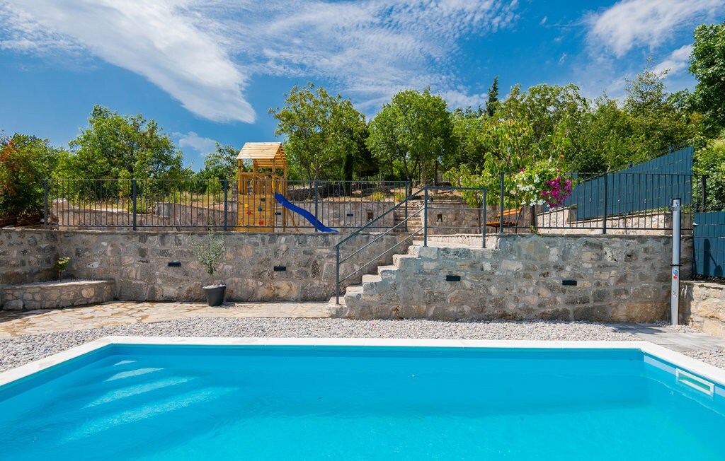An outdoor area features a clear blue swimming pool surrounded by natural stone walls. In the background, a children’s playground with a slide is visible, along with trees and flowering plants, adding a touch of greenery to the space.