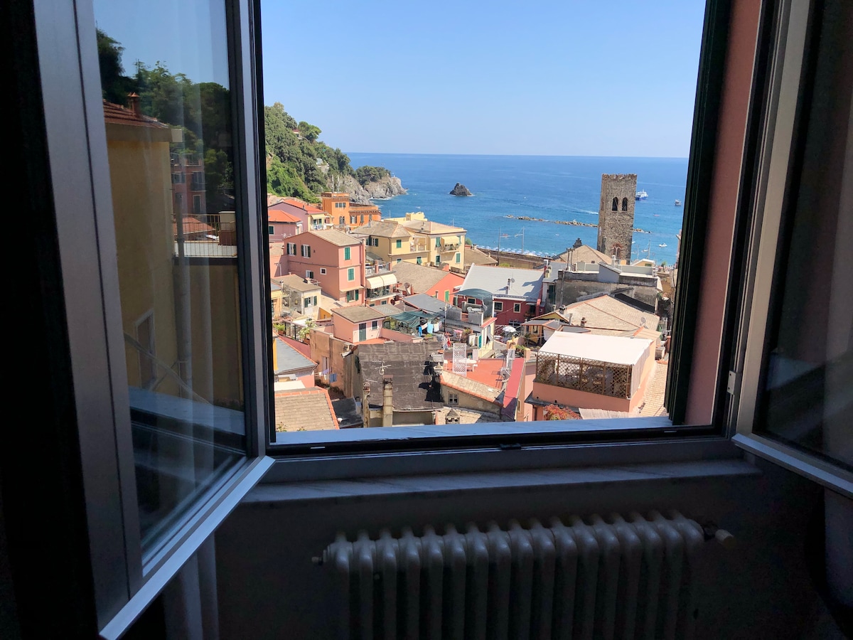 A view is offered through an open window, showcasing the vibrant rooftops of the town, the coastline with a rocky outcrop, and the distant ocean. A tower stands prominently amid the colorful buildings, framed by the window's edges.