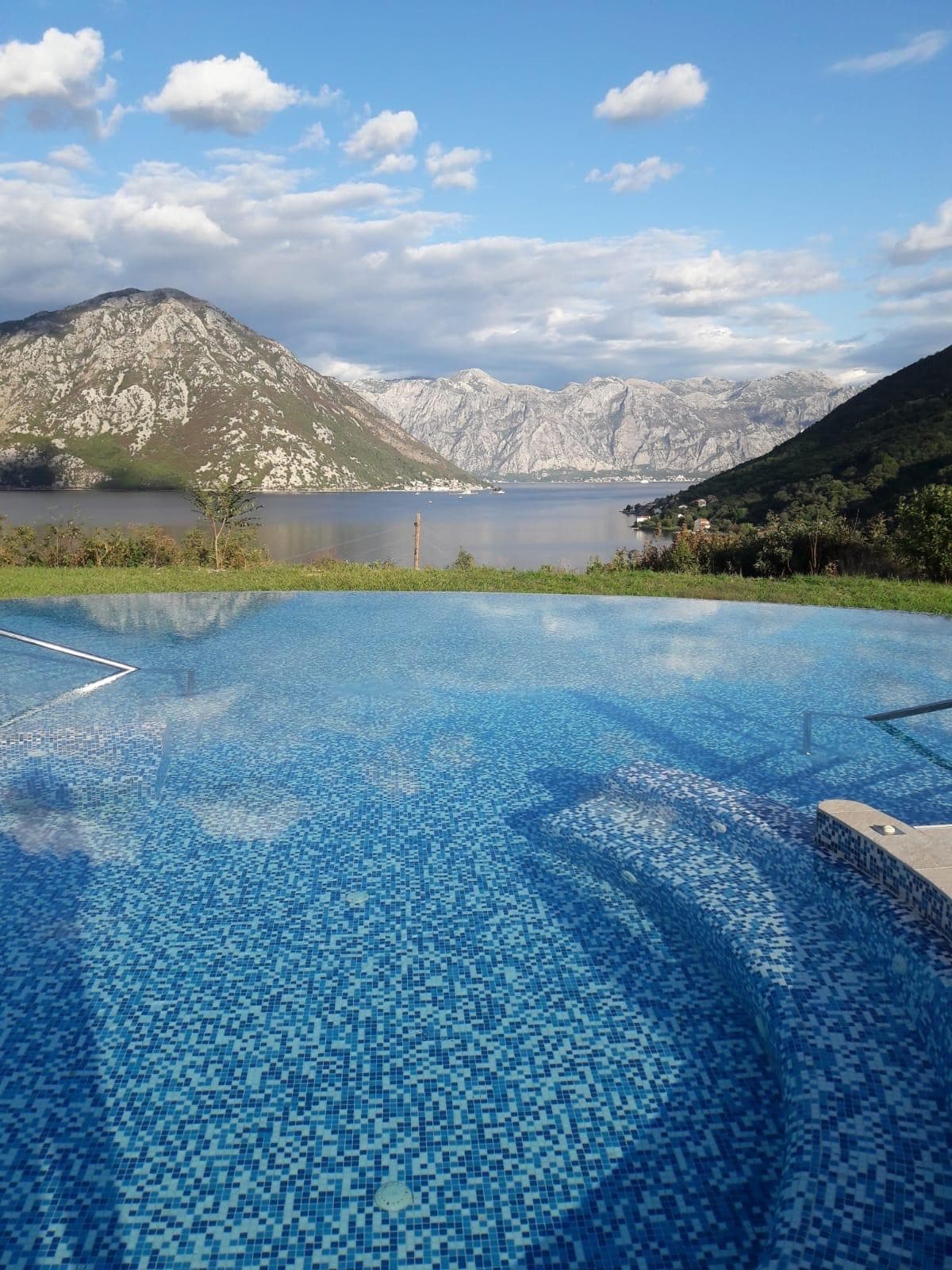 The image captures a serene pool overlooking a scenic landscape of mountains and water. The pool features mosaic tiles in shades of blue, reflecting the sky and surrounding nature. A tranquil atmosphere is created by the picturesque mountains in the background.
