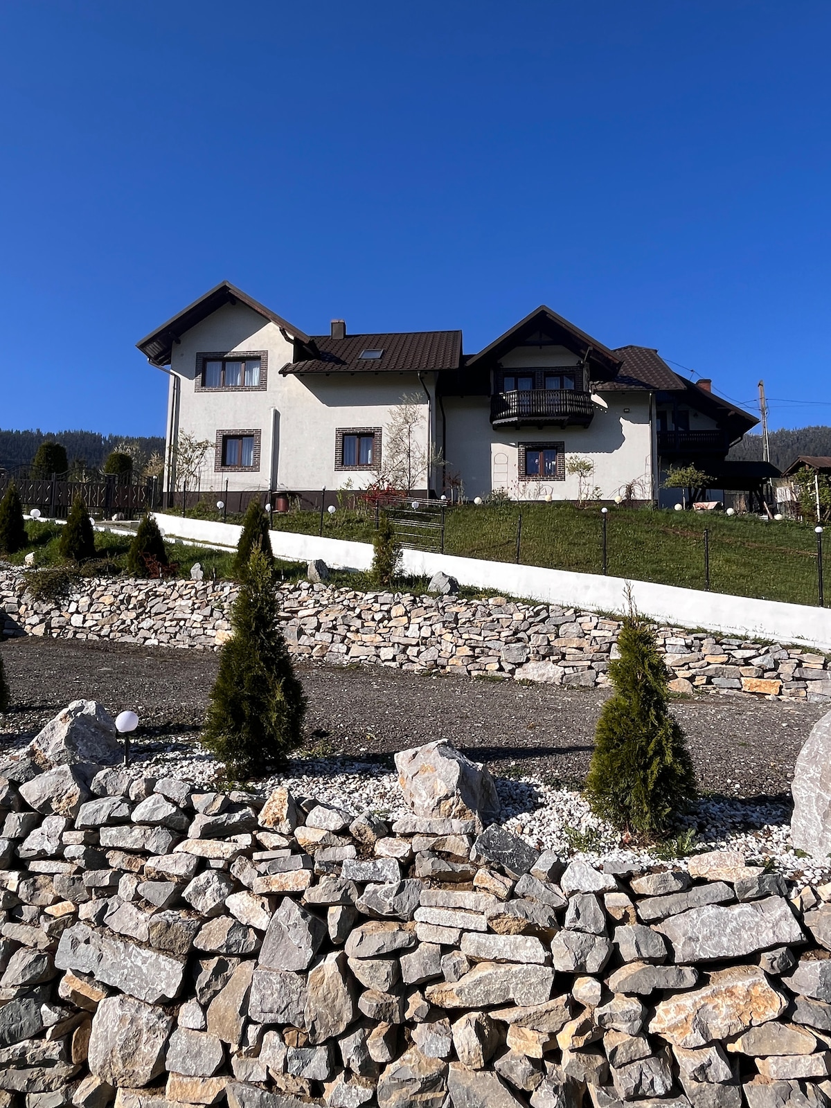 A charming multi-story house is set against a clear blue sky, surrounded by a well-maintained garden. Pathways lined with stones lead up to the residence, complemented by neatly trimmed shrubs and a lush lawn in the foreground.