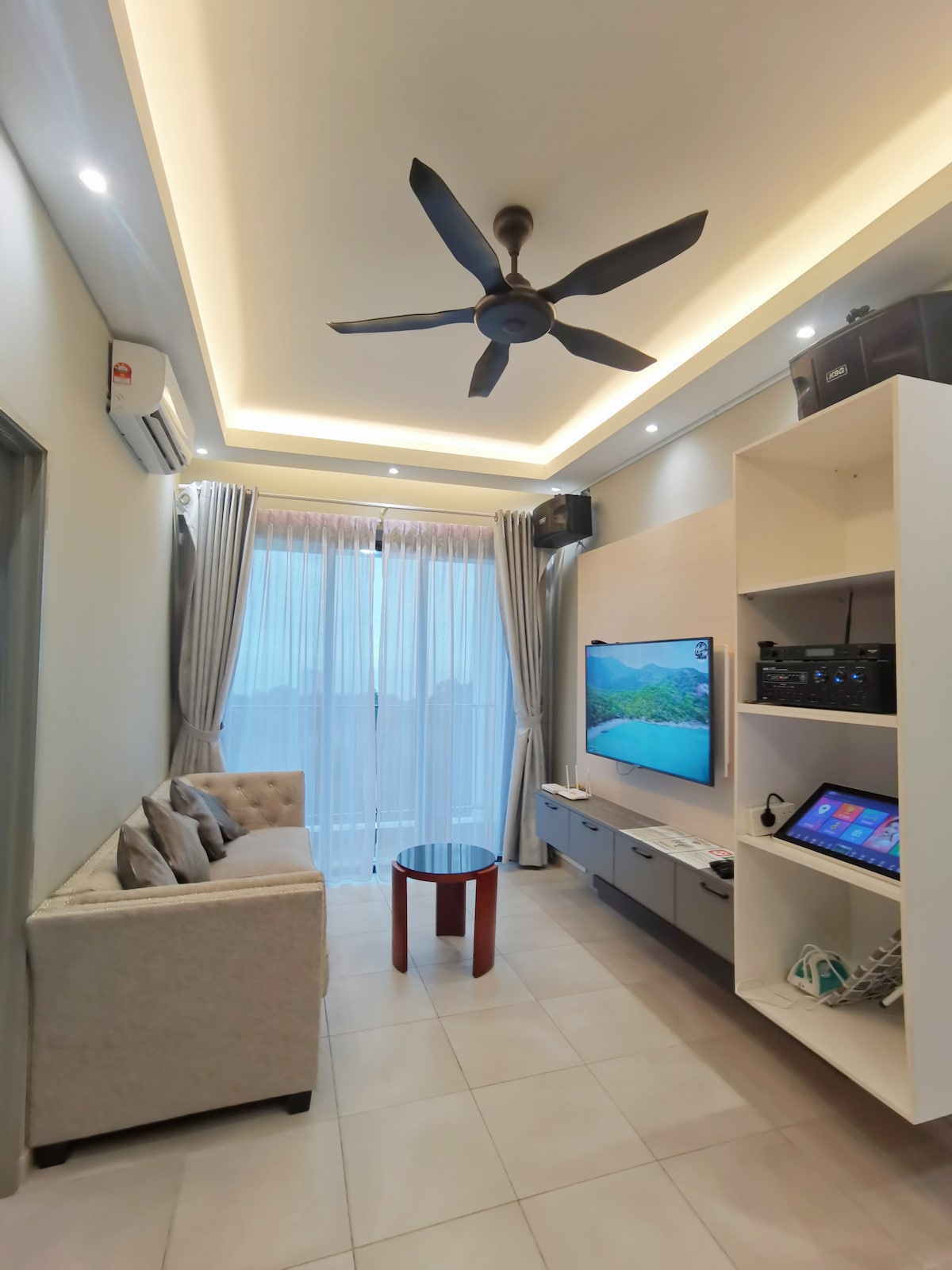 A comfortable living area is highlighted by a soft sofa and a small table, with a mounted television and entertainment system nearby. Tall windows are covered by light curtains, allowing natural light to fill the space, while a ceiling fan enhances air circulation.