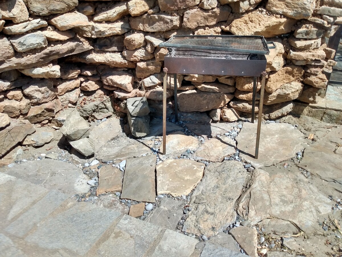 A simple outdoor grilling station is set against a rustic stone wall. The grill is metallic with a flat cooking surface, positioned on sturdy legs over a pathway made of natural stone slabs. Surrounding rocks add a natural touch to the area.