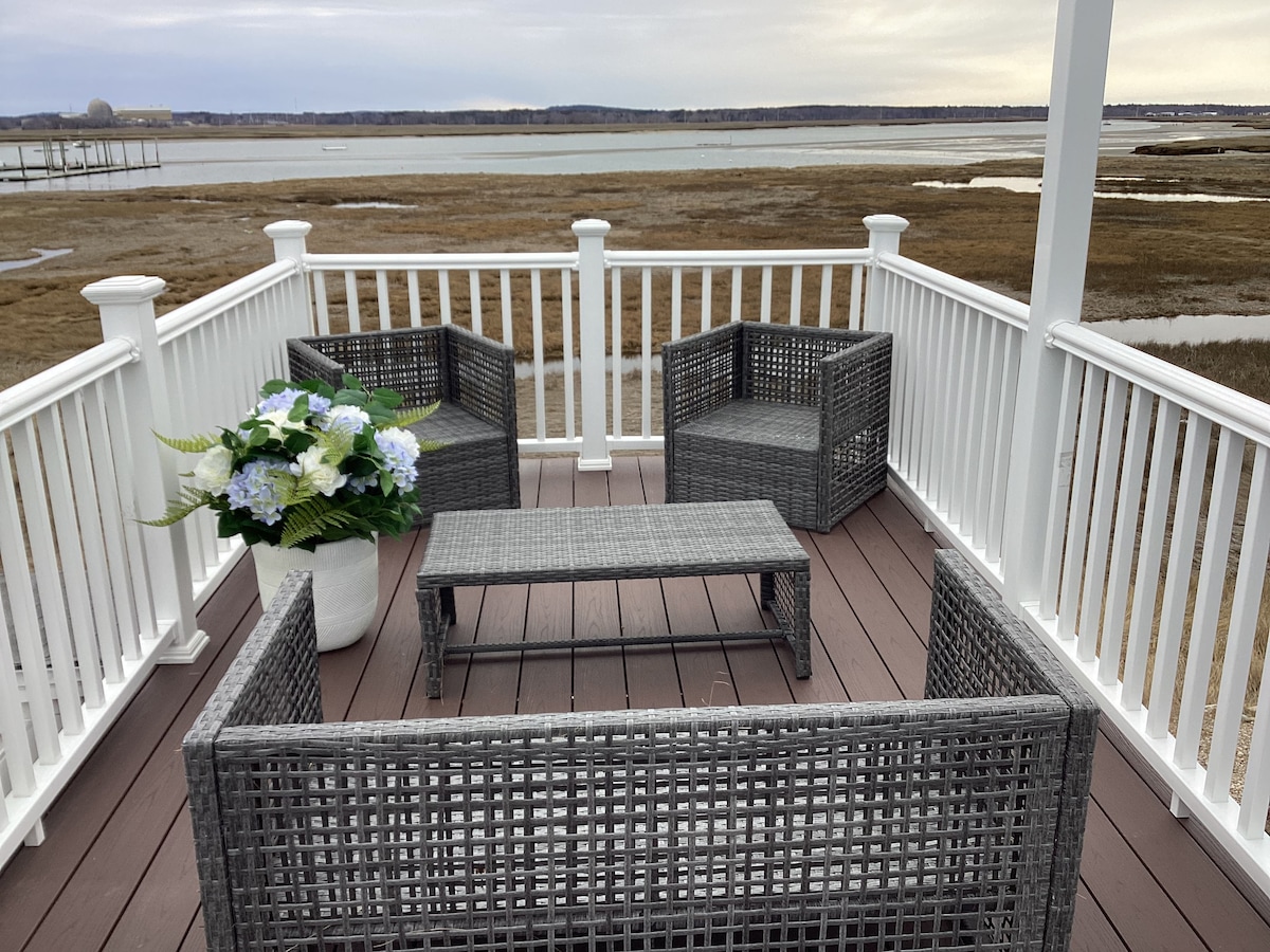 The rooftop deck features gray wicker seating arranged around a central table. A large white planter filled with blue and green flowers adds a pop of color. The view overlooks a tranquil river and expansive wetlands, creating a peaceful outdoor environment.