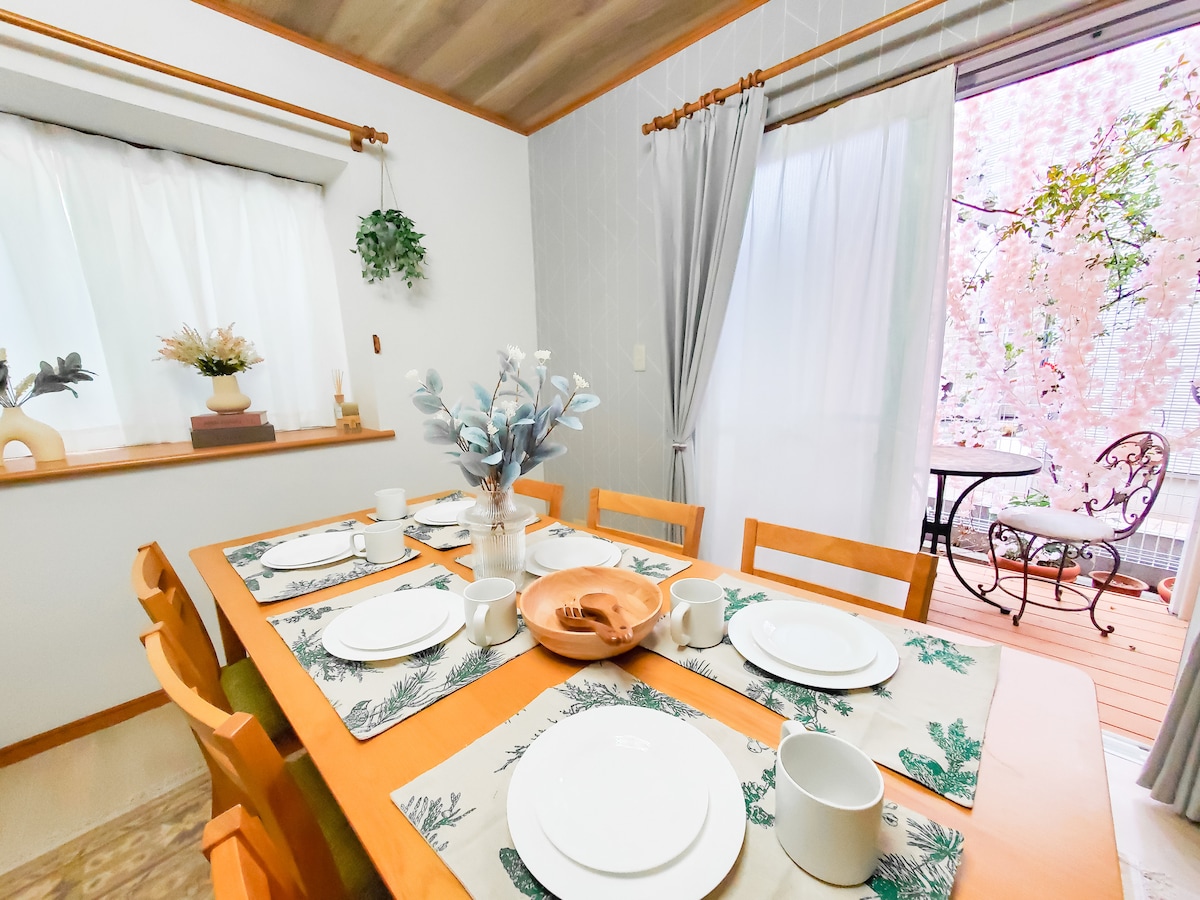 A dining area features a wooden table set with white dishes and cups, complemented by a floral centerpiece. Sunlight filters through sheer curtains, enhancing the inviting space. A doorway leads to an outdoor area with a small table and chairs, surrounded by greenery.
