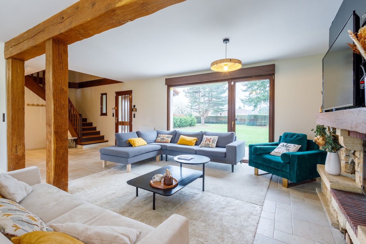 A spacious living area features two large sofas with cushions, a coffee table at the center, and a decorative rug underfoot. Natural light fills the room through large windows overlooking the garden. A TV is mounted on the wall, complemented by wooden beams and a staircase in the background.