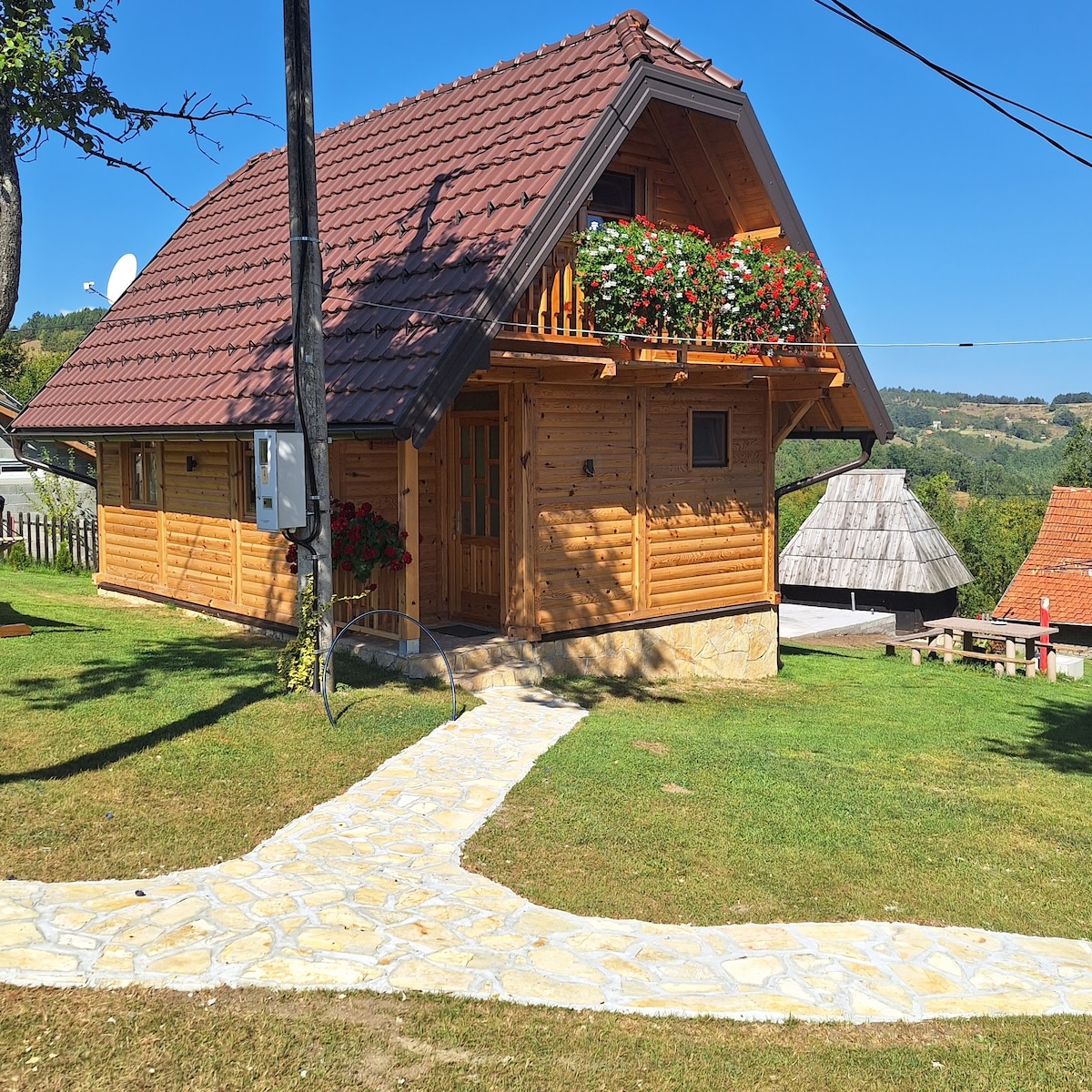 A wooden cabin is presented with a sloped roof and a charming balcony adorned with vibrant flower boxes. A stone pathway leads to the entrance, surrounded by a green lawn. Nearby structures and natural scenery enhance the peaceful rural setting.