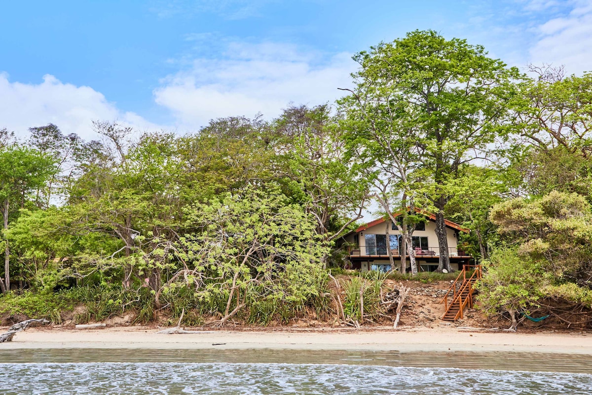 Surrounded by vibrant greenery, the beach house is set back from the shore, accessible by a wooden staircase. Lush trees provide a natural canopy, enhancing the serene coastal atmosphere. The structure features a simple architecture, with a balcony visible from the beach, inviting relaxation.