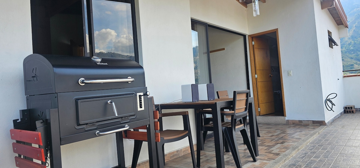 An outdoor balcony features a sturdy charcoal grill complemented by a table and chairs arranged for dining. Large sliding glass doors provide access to the interior, while scenic views of the mountains are visible in the background. The space is well-lit by natural daylight.