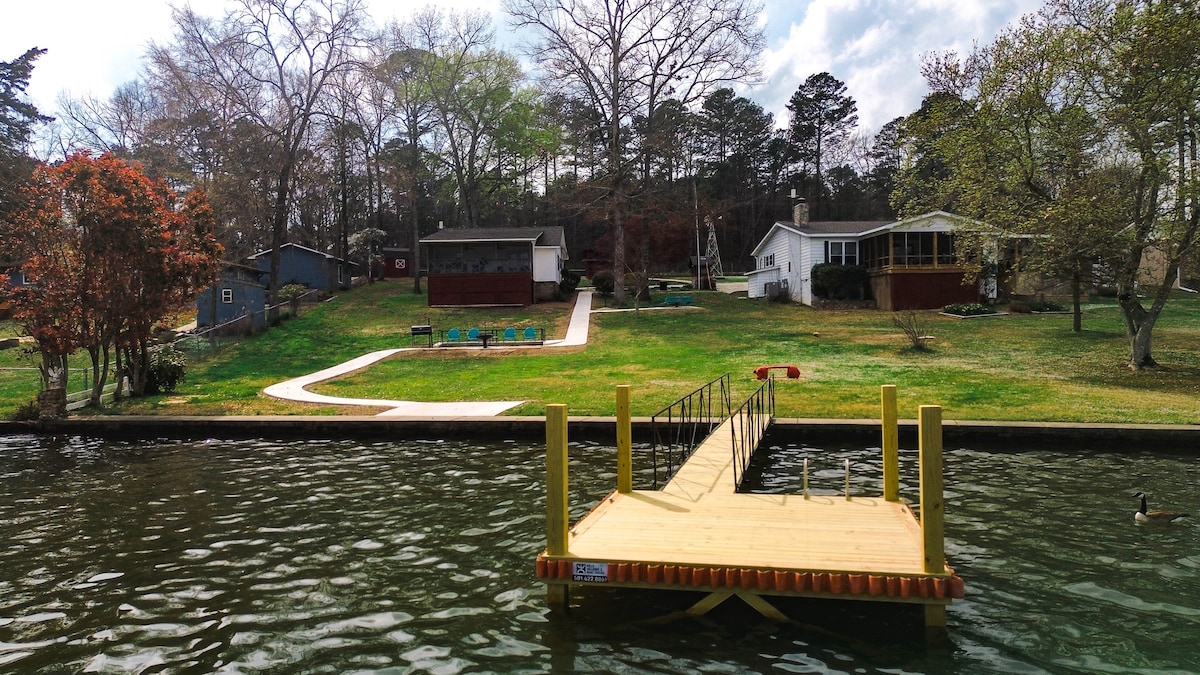 A private boat dock extends over the calm waters, providing direct access to Lake Hamilton. The surrounding yard showcases a spacious lawn with well-maintained greenery, leading to a charming cottage and a screened porch in the background. Trees and wooden accents enhance the natural setting.