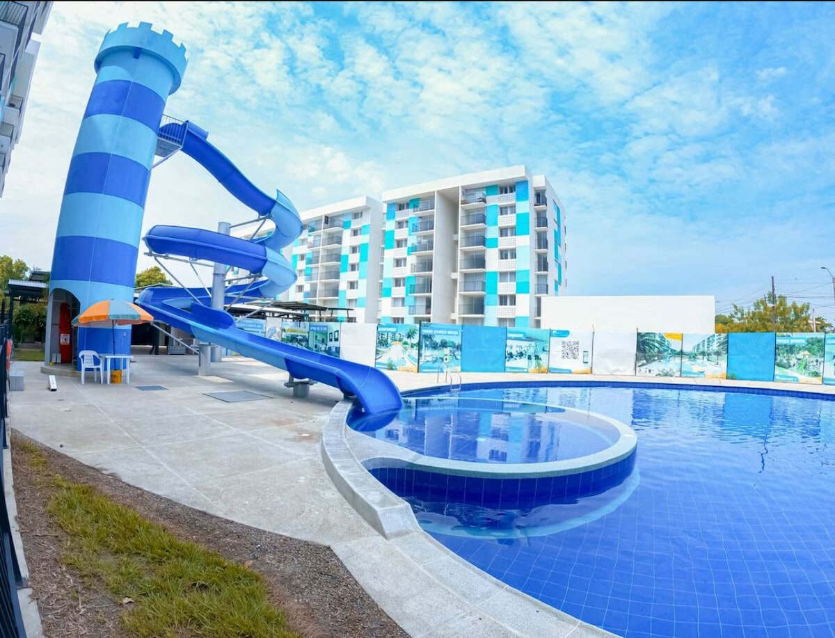 An outdoor pool area features a large circular pool surrounded by a spacious deck. A blue water slide curves from a tall, striped tower nearby. Colorful umbrellas provide shade, and a backdrop of a modern building enhances the recreational setting.