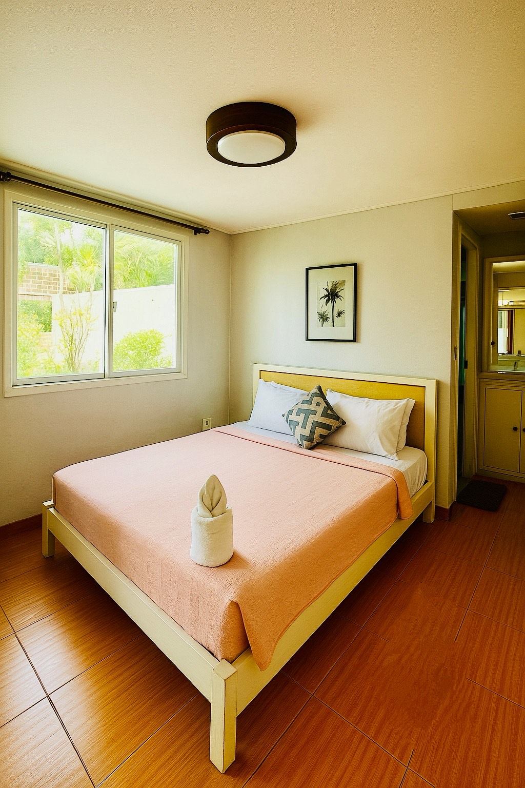 A standard room is presented with a comfortable queen bed topped with a patterned pillow and a light blanket. Natural light enters through a large window, illuminating the wooden floor. A tasteful wall art piece is displayed, contributing to the serene ambiance.