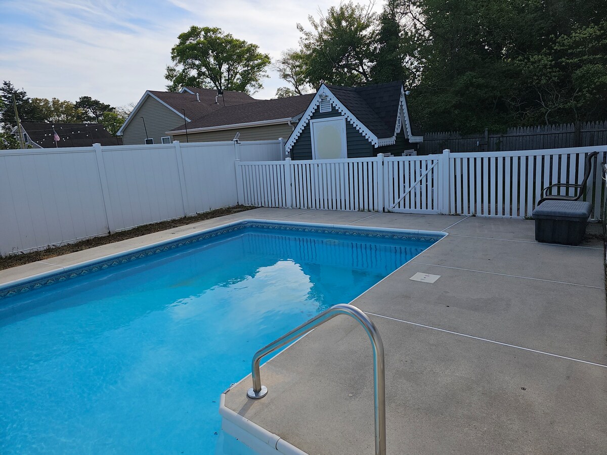 A private in-ground pool is surrounded by a white fence, featuring a concrete deck. The clear blue water reflects the sky, while a chaise lounge sits at the poolside. A nearby shed is visible against the backdrop of a residential area.