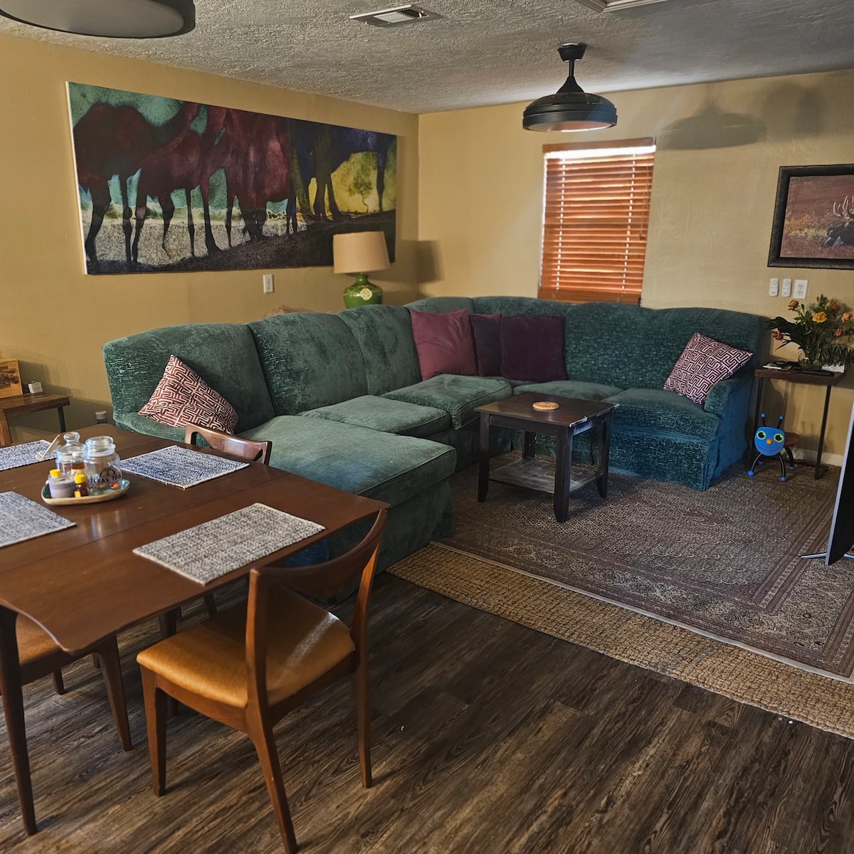 A spacious living area features a large, green sectional sofa with colorful accent pillows. A brown wooden table is set for dining, complemented by woven placemats. Artwork adorns the wall above, and natural light filters through the partially open window.